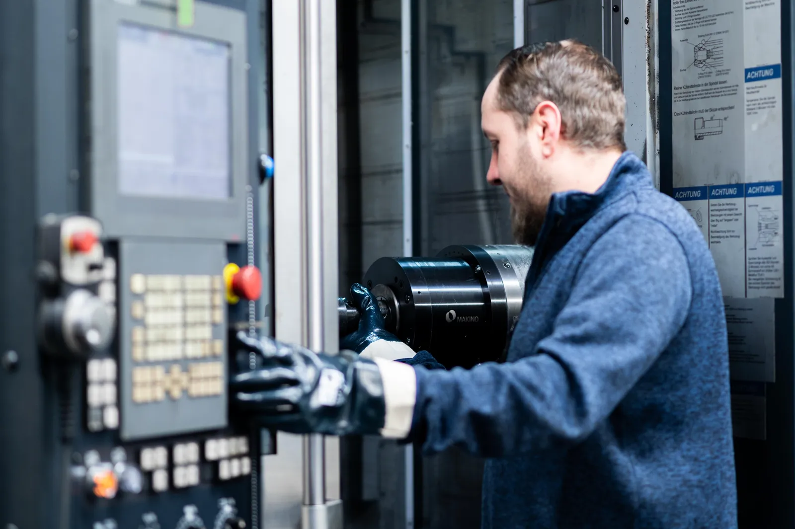 Mann mit blauer Jacke und Handschuhen bedient CNC-Maschine in industrieller Umgebung mit Warnhinweisen an der Wand.