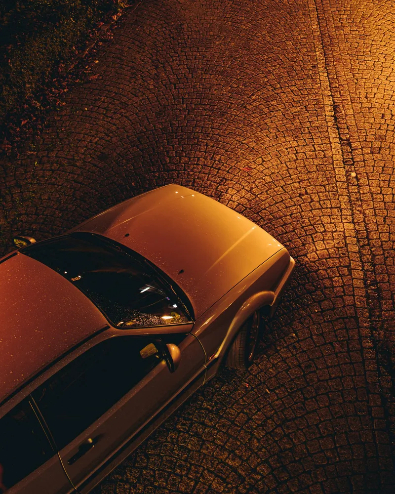 Teilansicht eines geparkten Autos auf nassem Kopfsteinpflaster bei Nacht mit warmem Straßenlicht.